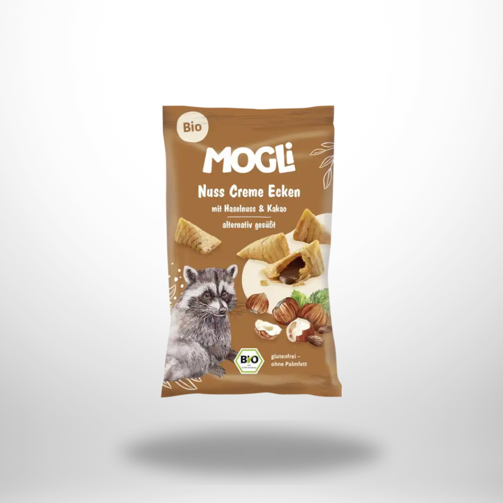 Brauner 30-g-Beutel „MOGLi Nuss Creme Ecken“ mit Haselnuss-Kakao-Füllung; einzelne Dreieck-Snacks und Haselnüsse daneben.